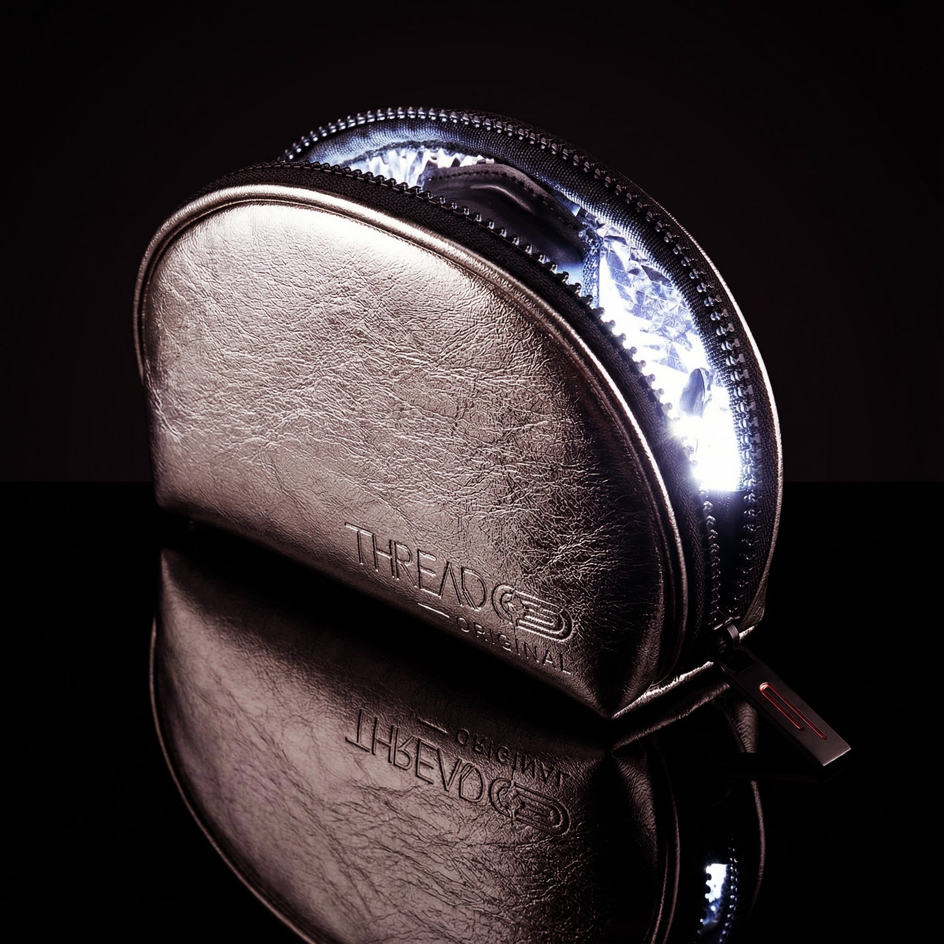 Champagne metallic LED makeup bag with illuminated interior on reflective black surface. Compact vegan leather cosmetic pouch with rechargeable light and wipeable insulated lining by Thread Original.