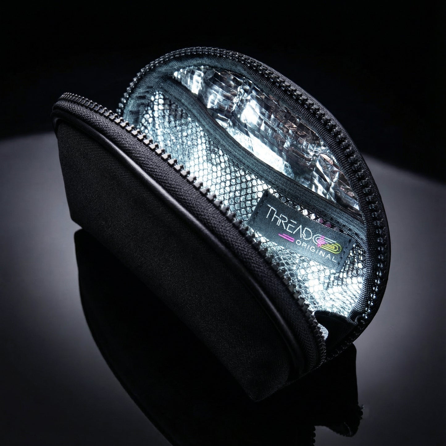 Black metallic LED makeup bag with illuminated interior on reflective black surface. Compact vegan leather cosmetic pouch with rechargeable light and wipeable insulated lining by Thread Original.
