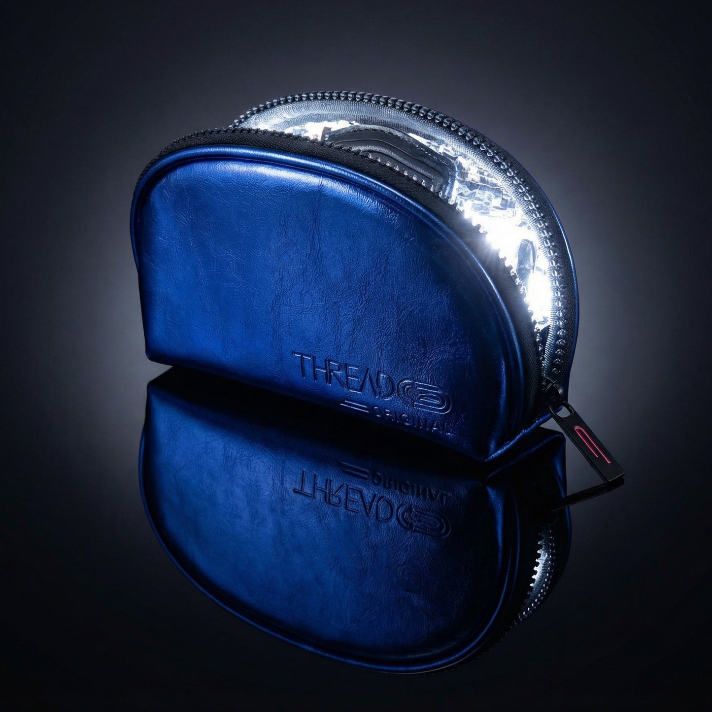 Ocean blue metallic LED makeup bag with illuminated interior on reflective black surface. Compact vegan leather cosmetic pouch with rechargeable light and wipeable insulated lining by Thread Original.