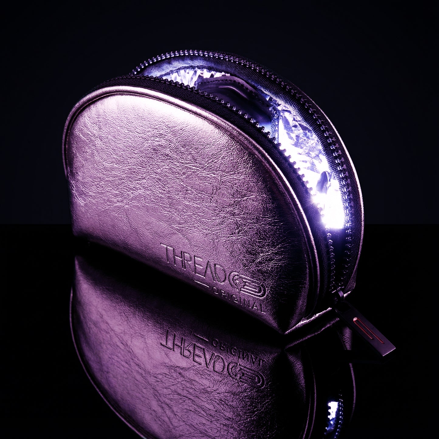 Pink metallic LED makeup bag with illuminated interior on reflective black surface. Compact vegan leather cosmetic pouch with rechargeable light and wipeable insulated lining by Thread Original.