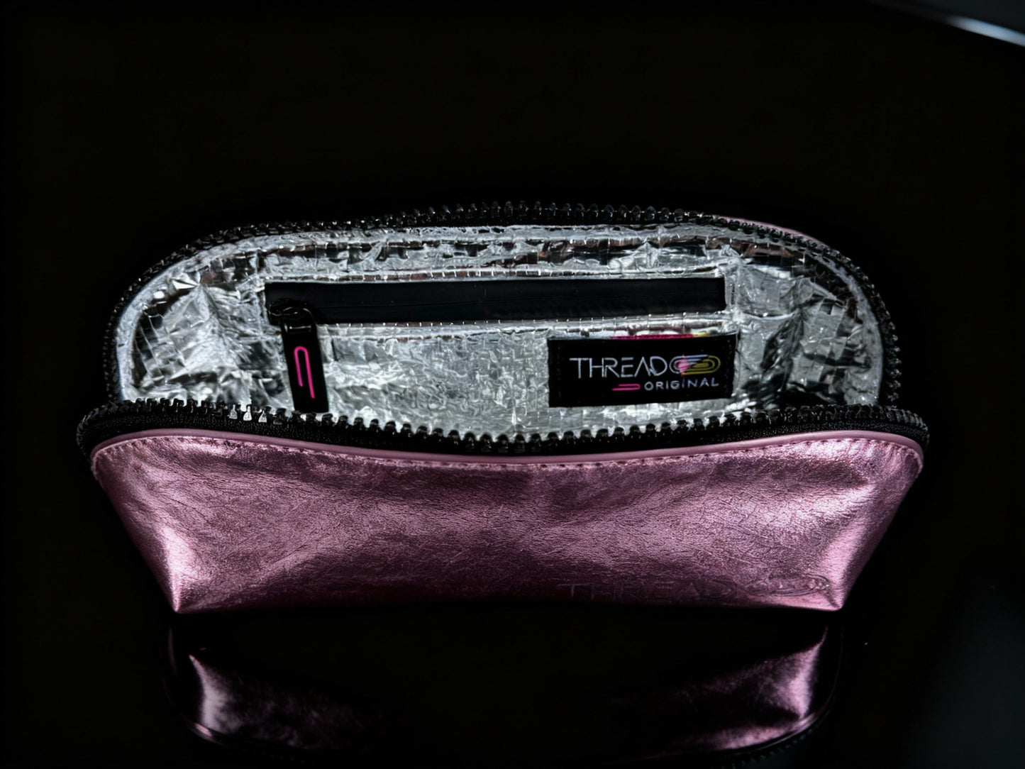 Pink metallic compact makeup bag with wipeable insulated interior on reflective black surface. Slim vegan leather cosmetic pouch by Thread Original.