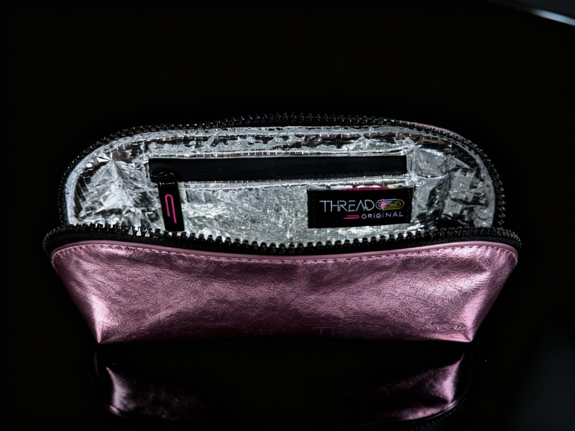 Pink metallic compact makeup bag with wipeable insulated interior on reflective black surface. Slim vegan leather cosmetic pouch by Thread Original.
